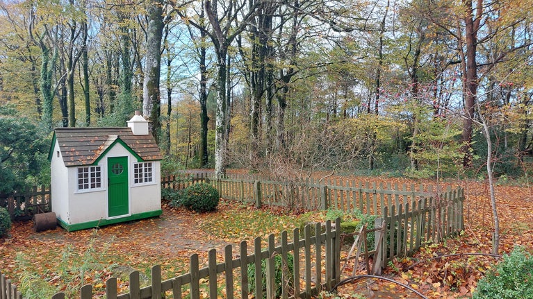 Children's play house with white walls and a green door. There is a small garden in front of the house and a fence.